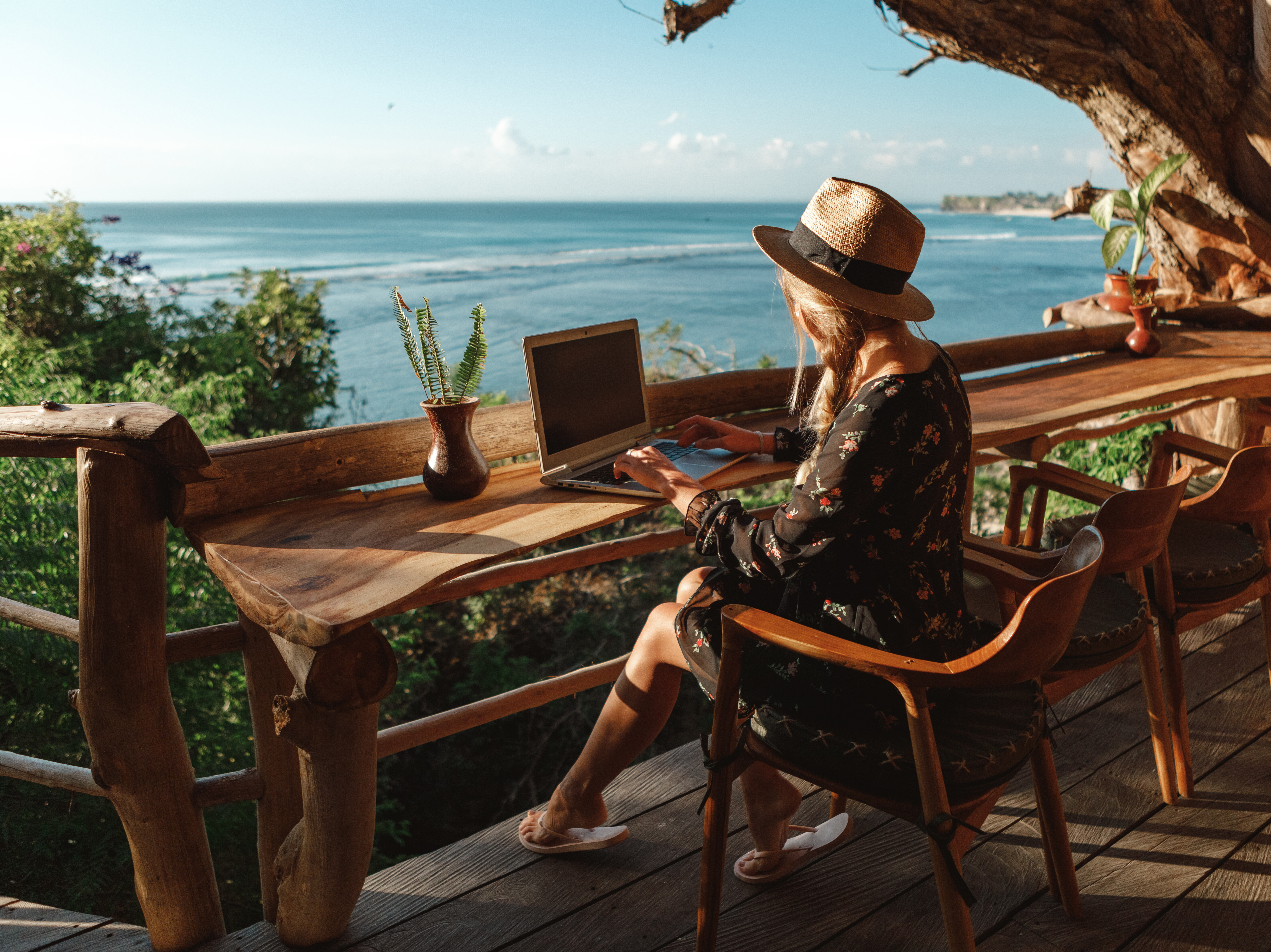 Frau sitzt mit Strohhut an einem Laptop auf einer Terrasse am Meer.